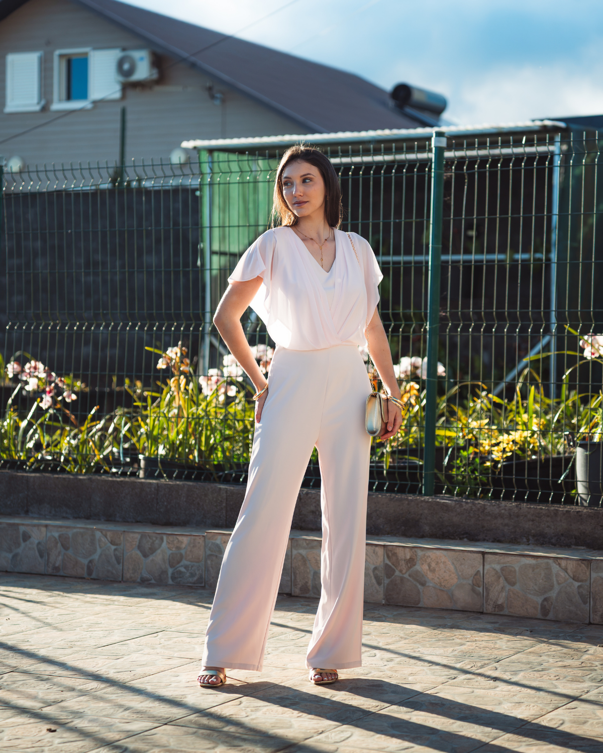Femme portant une combinaison pantalon rose poudré à haut cache-cœur voilé et jambes larges, avec une pochette dorée