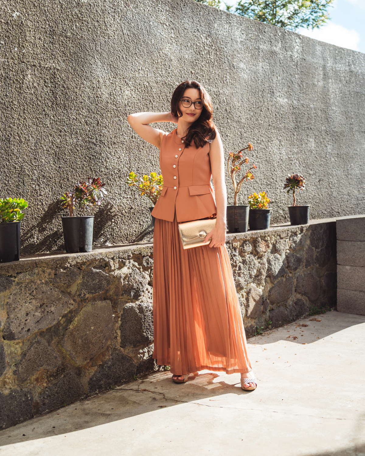 Femme portant un ensemble terracotta composé d’un gilet sans manches à boutons nacrés et d’une longue jupe plissée, avec une pochette dorée